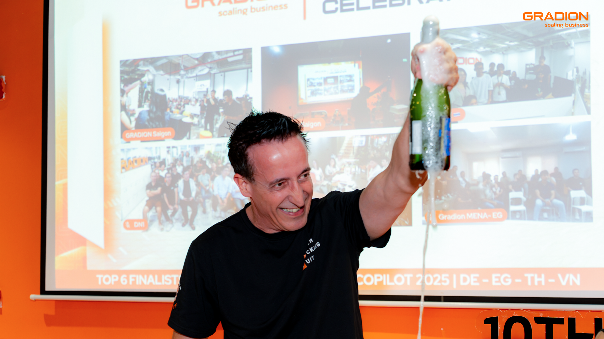 Gradion team celebration for 10th anniversary, with a leader raising a champagne bottle in front of a presentation screen showing highlights of company milestones.